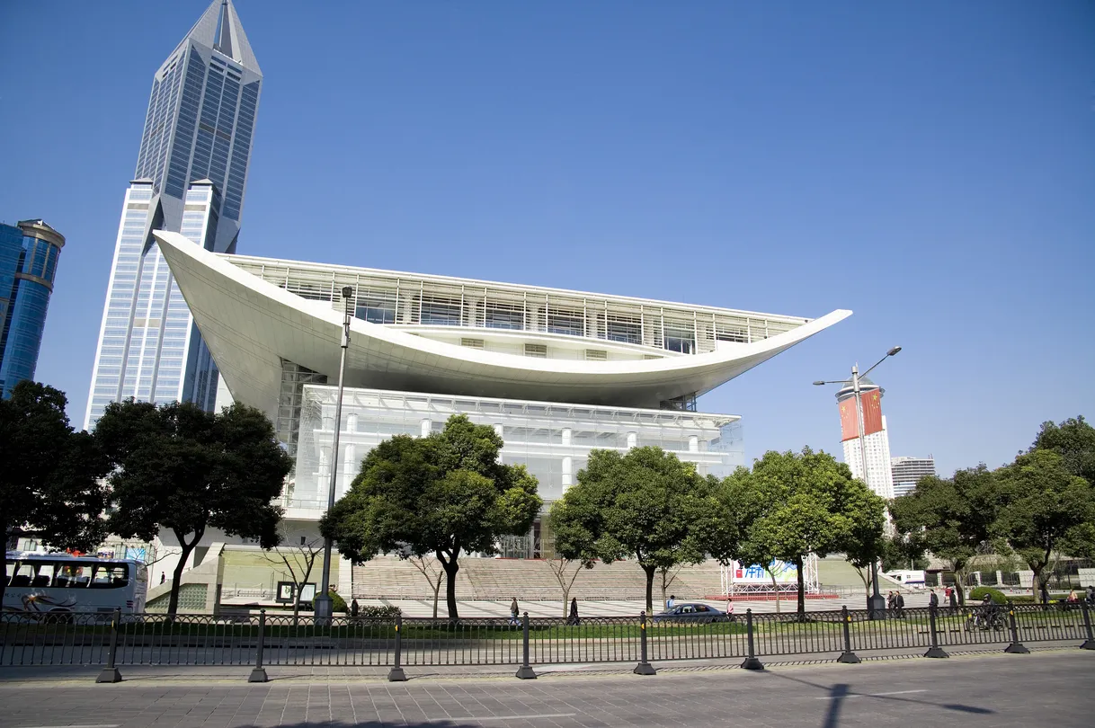 上海大劇院の建物外観。青空の下、屋根が翼のように広がる白いモダンな建築がそびえ、背後には高層ビルが立ち並ぶ都会的な風景。