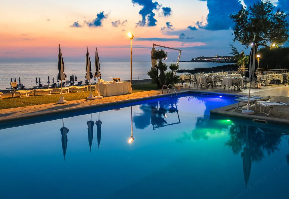 a beautiful outdoor swimming pool area with clear blue water , surrounded by umbrellas , and lit up at night at Hotel Poseidon