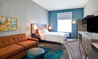 a modern hotel room with blue walls , white bedding , and wooden floors , featuring a bed , couch , coffee table , and window at Home2 Suites by Hilton Mechanicsburg