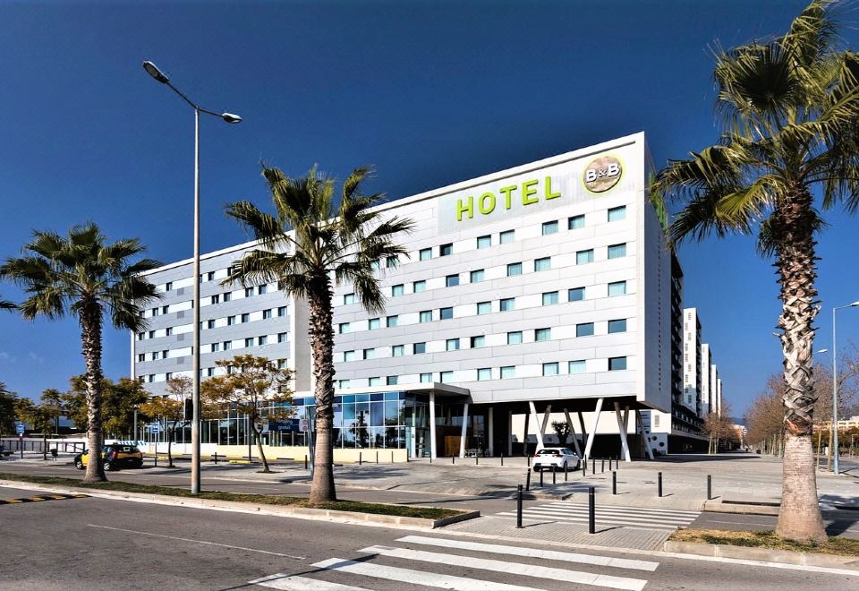 "a large hotel building with the name "" hotel "" written on it , surrounded by palm trees and a street" at B&B Hotel Barcelona Viladecans