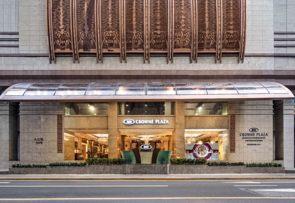 The front entrance of a hotel is depicted, featuring an illuminated sign above it, with other buildings visible in the background at Crowne Plaza Shanghai Nanjing Road