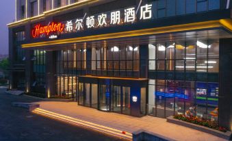 At night, a hotel entrance is adorned with an illuminated sign above its glass door at Hampton by Hilton Qingdao West Coast