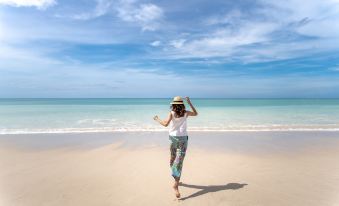 a woman in a hat and floral pants is standing on a beach with her arms raised at Grand Mercure Khao Lak Bangsak