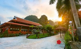 a beautiful sunset view of a traditional thai - style building with red flowers in front and palm trees in the background at Tam Coc La Montagne Resort & Spa