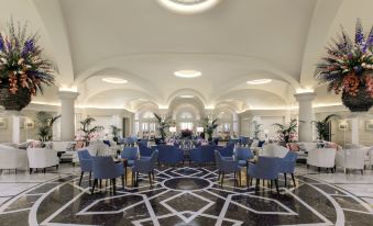 a large dining room with blue chairs and tables , surrounded by white columns and black and white checkered flooring at The Phoenicia Malta