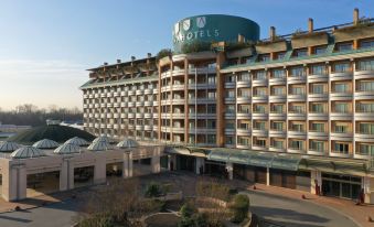 "a large hotel with a green sign that reads "" hotel "" is surrounded by trees and other buildings" at UNA Hotels Expo Fiera Milano