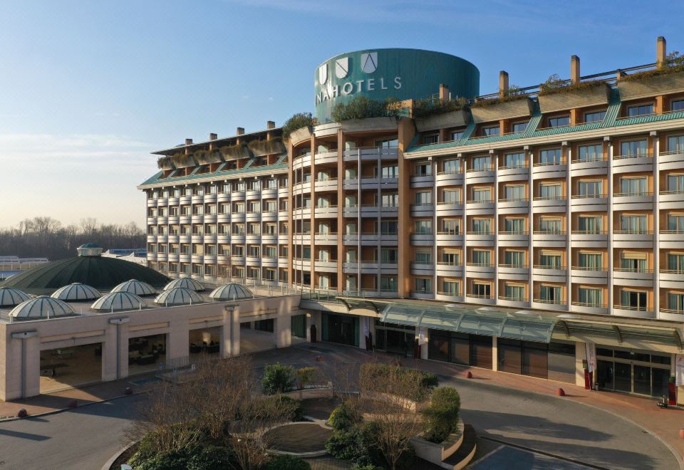 "a large hotel with a green sign that reads "" hotel "" is surrounded by trees and other buildings" at Unahotels Expo Fiera Milano