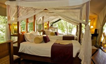 a cozy bedroom with two beds , one of which is covered by a canopy , and a view of trees outside through large windows at Mara Intrepids Tented Camp