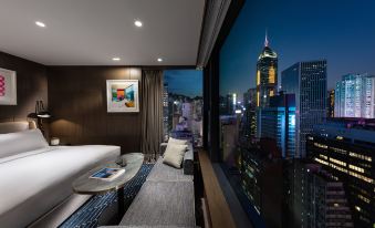 A bedroom with large windows overlooking the city is illuminated by a bed on one side at The Hari Hong Kong
