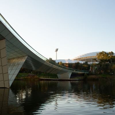 a modern , curved bridge with glass walls and a railing , reflecting in the calm water below at Oval Hotel at Adelaide Oval, an EVT hotel