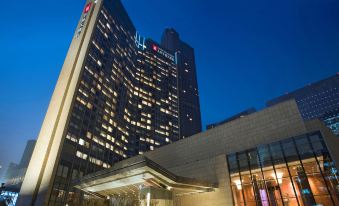 At night, there is a large building in front of the hotel with an illuminated sign on top at Grand Millennium Beijing