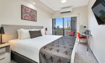 a large bed with a white headboard and gray blanket is in a room with a desk , chair , and sliding glass doors leading to a at Argus Hotel Darwin