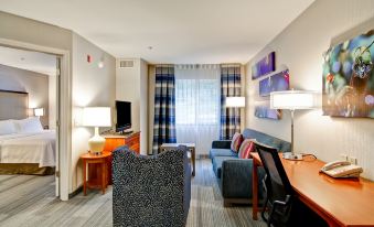 a modern hotel room with a blue and black patterned couch , desk , chairs , lamps , and a window with curtains at Homewood Suites by Hilton Stratford