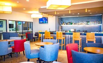 a modern bar with blue and yellow chairs , a television screen , and an array of bottles on the counter at Highlander Hotel & Apartments
