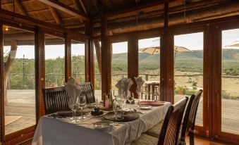 a dining table is set with wine glasses and a plate of food , surrounded by chairs , in front of a scenic view at Pumba Private Game Reserve