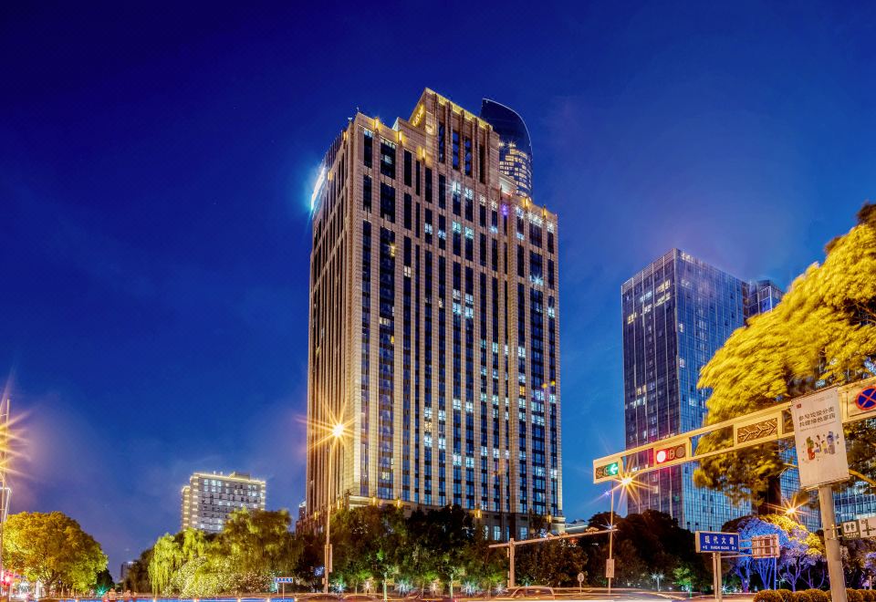 a tall building with a blue sky in the background , surrounded by other buildings and trees at Suisse Apartment Hotel Suzhou Jinji Lake Expo Center