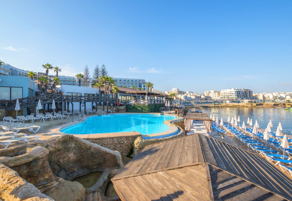 a large outdoor pool surrounded by lounge chairs and umbrellas , with a view of the ocean in the background at DoubleTree by Hilton Malta