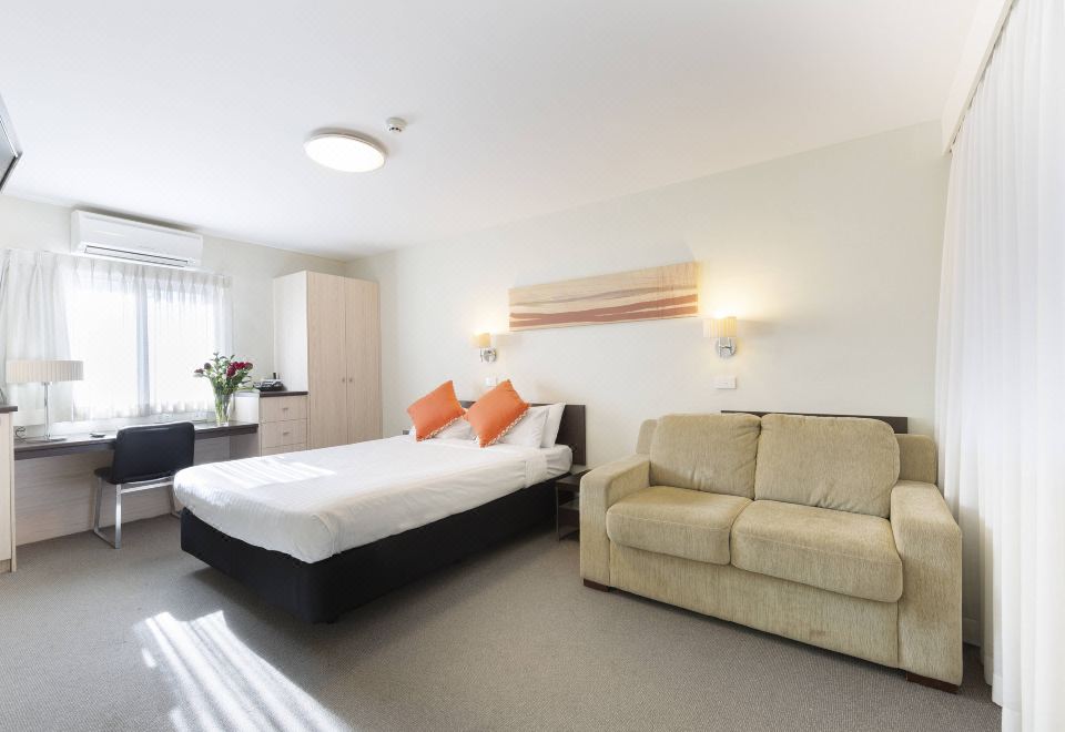 a modern hotel room with a large bed , white bedding , and an orange throw pillow , along with a beige couch and a window at Ibis Styles Canberra Tall Trees