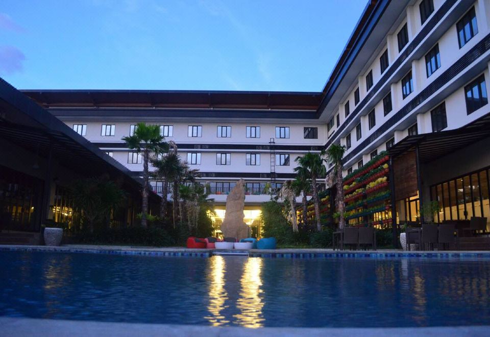 a large swimming pool is surrounded by tall buildings and palm trees , with the sun casting a warm glow on the water at Hotel Neo Eltari - Kupang by Aston