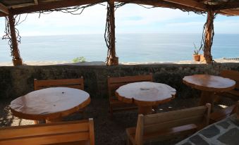 a restaurant with wooden tables and chairs , overlooking the ocean , under a thatched roof with trees and water visible in the background at Antulang Beach Resort