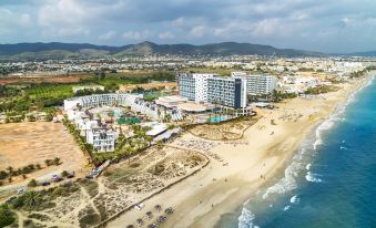 aerial view of a sandy beach with a hotel in the distance , surrounded by palm trees and a body of water at Hard Rock Hotel Ibiza