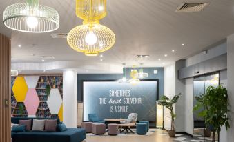 "a modern office lobby with a large screen displaying "" sometimes the best souvenir is a smile "" and various furniture pieces" at Hampton by Hilton Rome North Fiano Romano