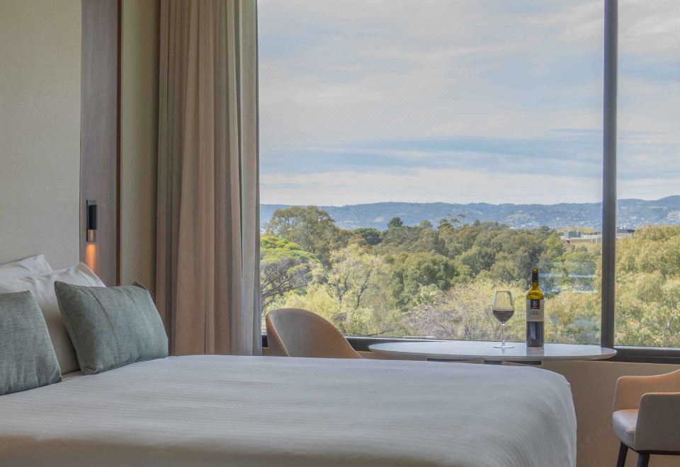 a bed with a white sheet and pillow , next to a window that offers a view of trees and mountains at Oval Hotel at Adelaide Oval, an EVT hotel