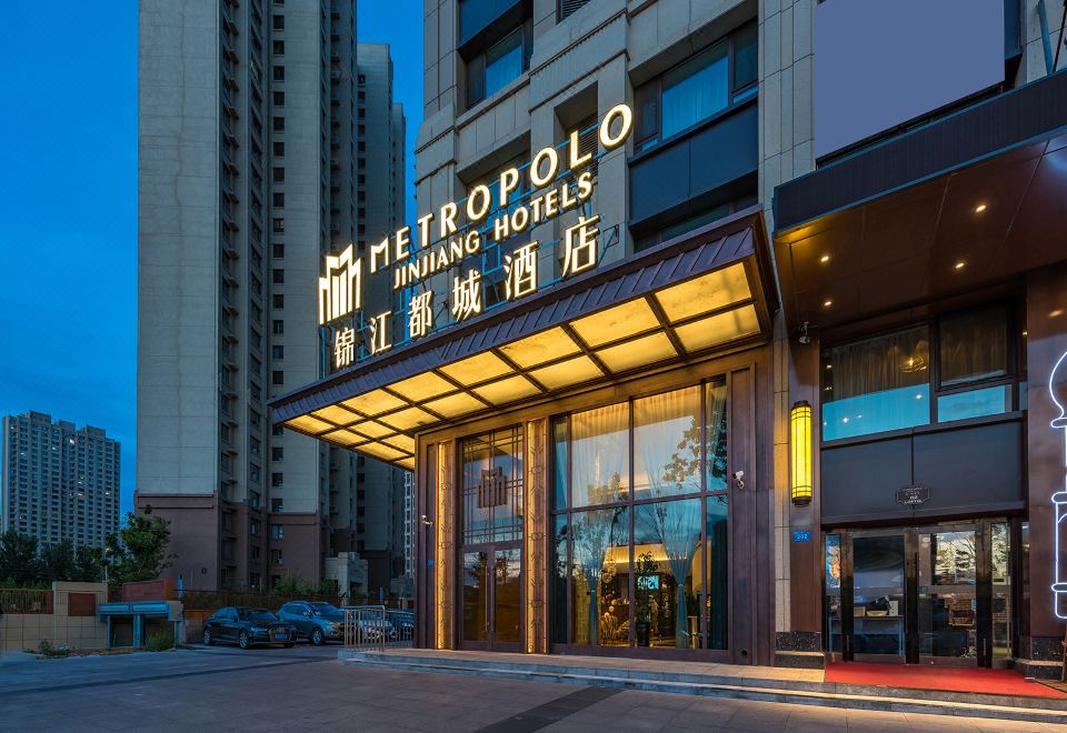 At night, a hotel's front entrance is illuminated by a sign, surrounded by other buildings at Metropolo Jinjiang Hotel (Harbin Haxi High-speed Railway Station Wanda Plaza)