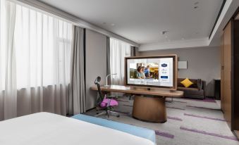 A room with large windows, a bed, and a desk is shown next to the television at Hilton Hampton Hotel, West Coast, Qingdao