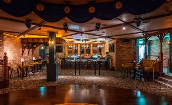 a dimly lit bar with wooden tables and chairs , surrounded by a bar counter and several potted plants at The Prince of Wales Hotel