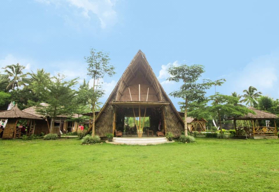 a large wooden building with a triangular roof is surrounded by green grass and trees at Balkondes Wringinputih