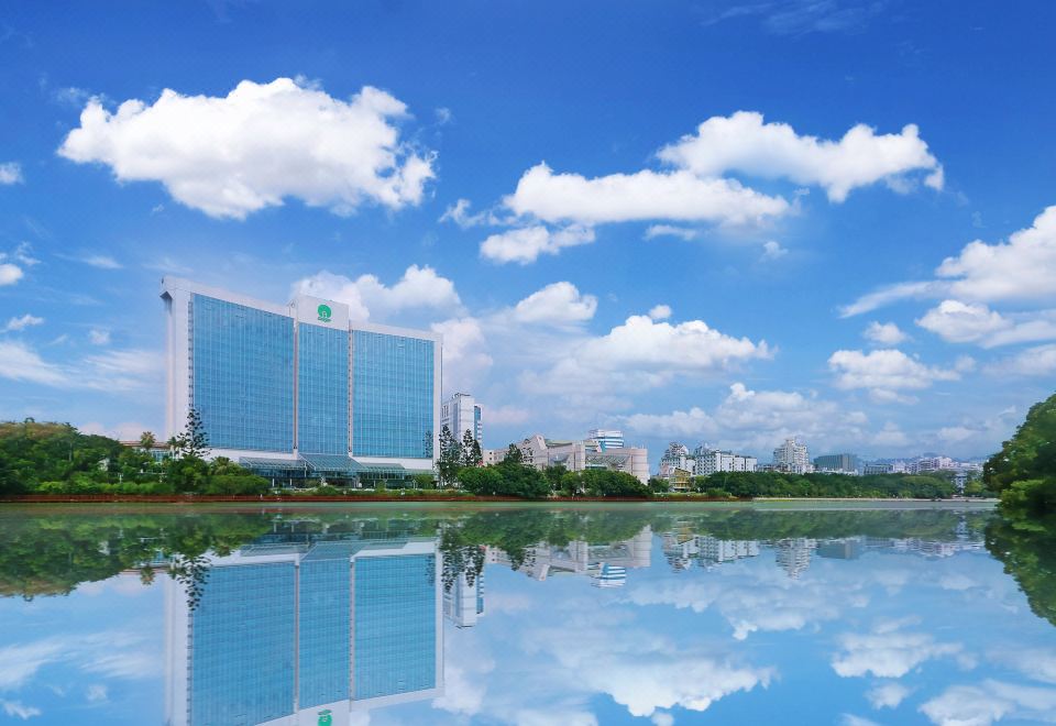 a large building with a blue glass facade is reflected in the water , surrounded by greenery and clouds at Lakeside Hotel