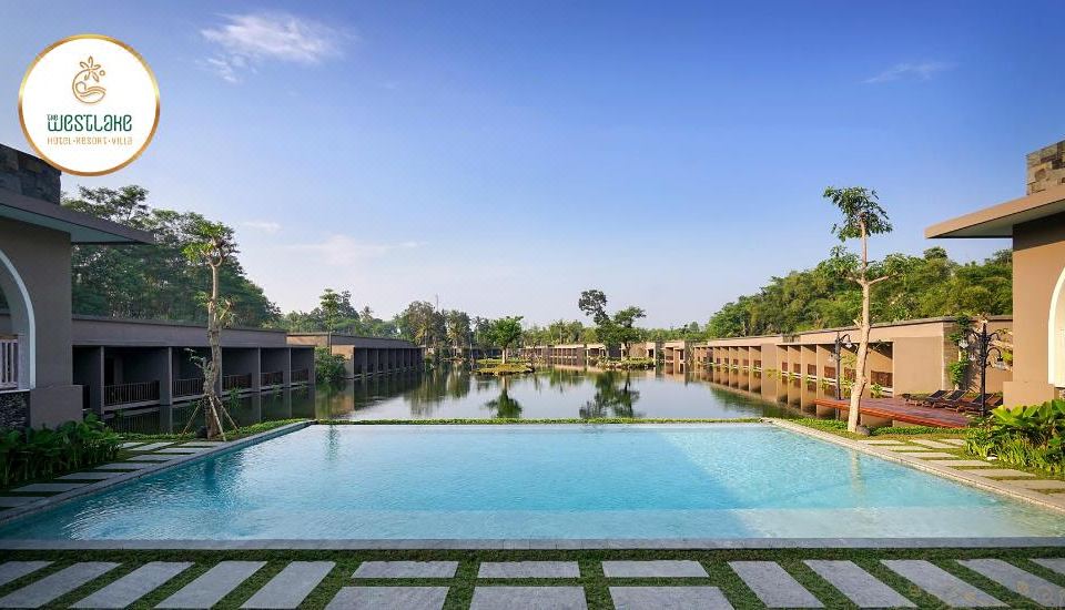 a large swimming pool is surrounded by a building and trees , with a clear blue sky above at The Westlake Hotel & Resort Yogyakarta