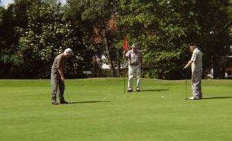 three men are playing golf on a green field with trees in the background , and one man is lining up a putt at Coulsdon Manor Hotel and Golf Club