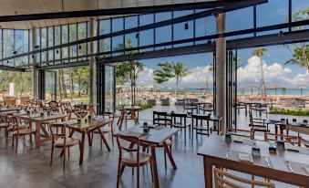 a large restaurant with wooden tables and chairs , set against a backdrop of palm trees and the ocean at La Vela Khao Lak