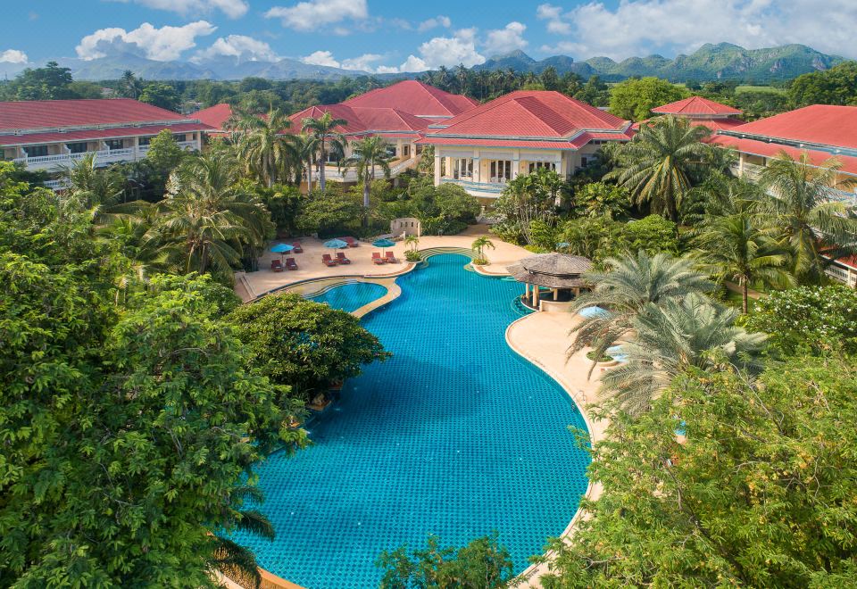 a large outdoor pool surrounded by lush greenery , with several lounge chairs and umbrellas placed around it at Dheva Mantra Resort