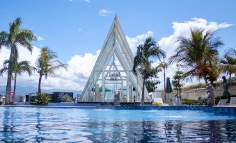 a large pool surrounded by palm trees , with a glass structure in the background and a building in the foreground at Swiss-Belhotel Silae Palu