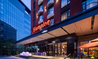 At night, a hotel's front entrance is illuminated by a sign above it, surrounded by other buildings at Hampton by Hilton Beijing Guomao CBD