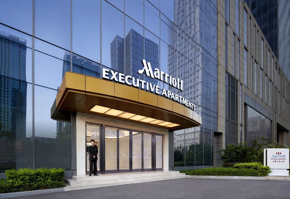 A hotel's front entrance is depicted with an illuminated sign above it, while another building can be seen in the background at the International Trade City, Yiwu - Marriott Executive Apartments