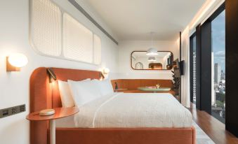The bedroom is illuminated by overhead lighting and features large windows and an orange accent wall above the bed at The Standard, Bangkok Mahanakhon