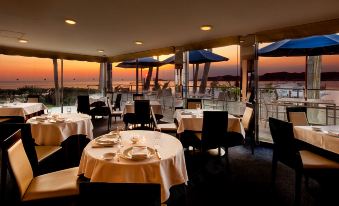 a large , well - lit restaurant with multiple dining tables and chairs , all set for a meal , under a sunset view at Malibu Hotel