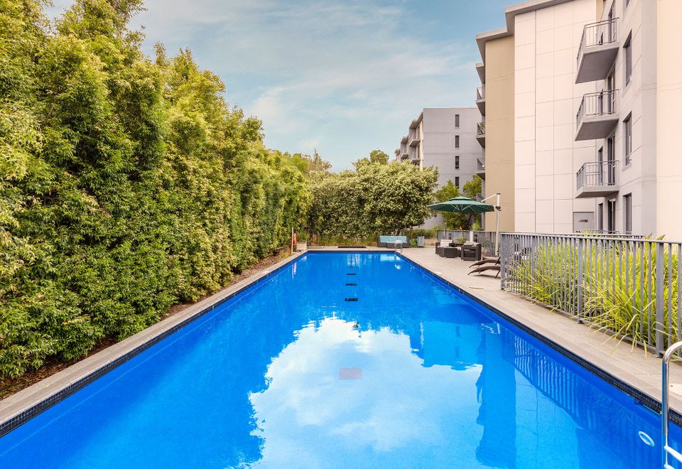 a large outdoor swimming pool surrounded by lush greenery , with a house in the background at Oaks Sydney North Ryde Suites