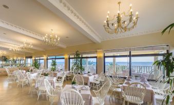 a large dining room with white tables and chairs , chandeliers hanging from the ceiling , and a view of the ocean at Potidea Palace Hotel