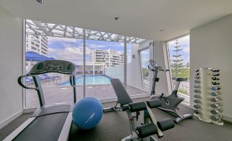 a gym with a blue exercise ball , exercise bike , and various exercise equipment is shown with a view of a swimming pool outside at The Sebel Mandurah