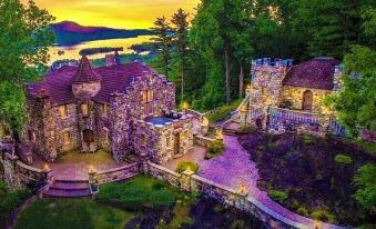 a large stone house surrounded by trees and a body of water , with a beautiful sunset in the background at Highlands Castle Overlooking Lake George Plus 2 Other Castles & Suites