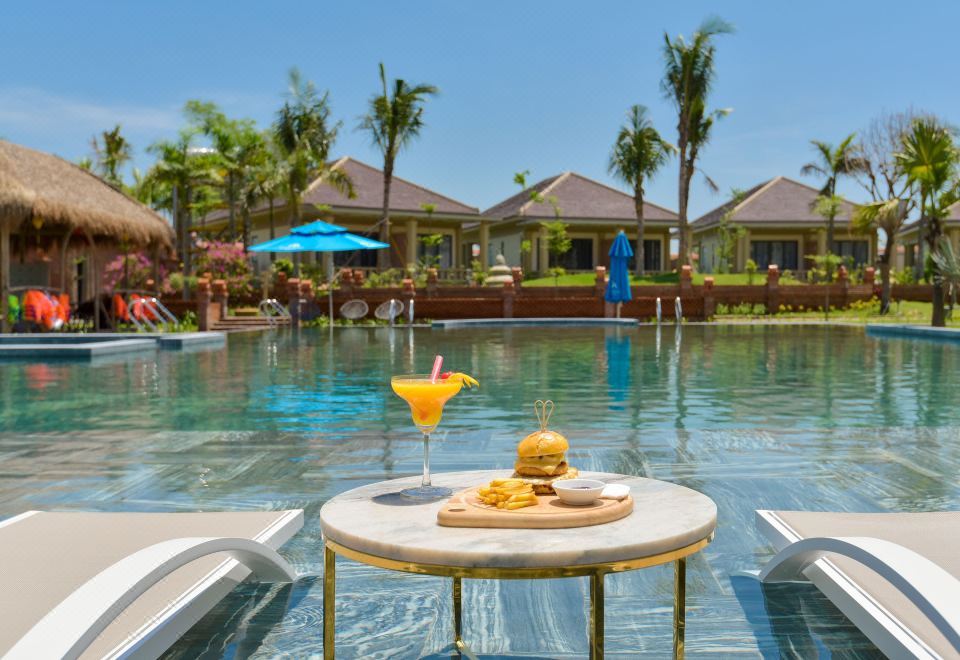 a poolside setting with a table with a drink and food items , surrounded by lounge chairs and umbrellas at My Son Heritage Resort and Spa