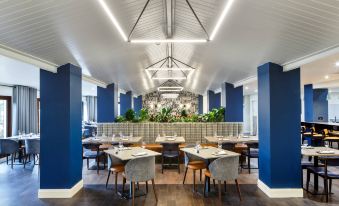 a modern restaurant with blue walls , wooden tables , and chairs , a bar area , and green plants at Delta Hotels Milton Keynes