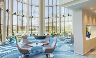 a modern , well - lit lounge area with large windows and blue carpeting , featuring white and gray chairs and tables at Hyatt Regency Long Beach