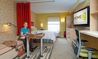 "a woman is sitting at a table in a hotel room with a large screen displaying "" home "" and red curtains" at Home2 Suites by Hilton Oxford