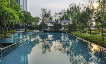 a long , empty swimming pool surrounded by lush greenery , with several lounge chairs placed around the pool at Swissôtel Jakarta PIK Avenue